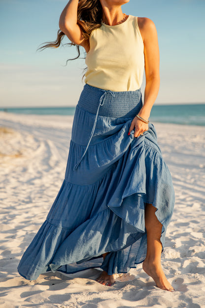 Woman in a yellow top and blue denim skirt standing on a sandy beach.