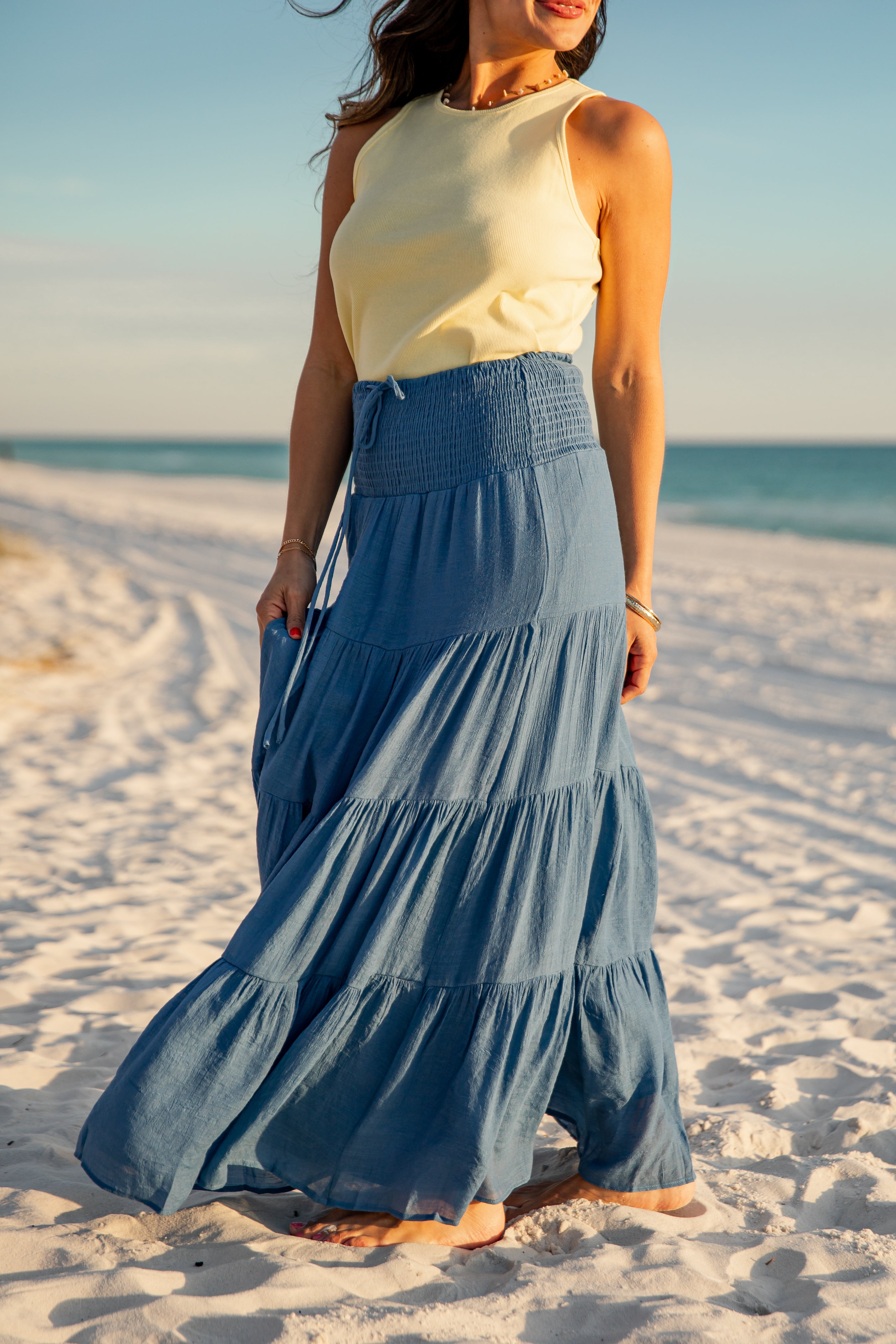 Woman in a yellow top and blue tiered skirt standing on a sandy beach.