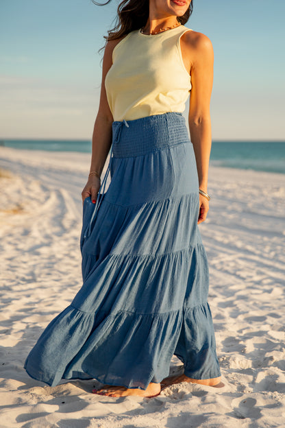 Woman in a yellow top and blue tiered skirt standing on a sandy beach.