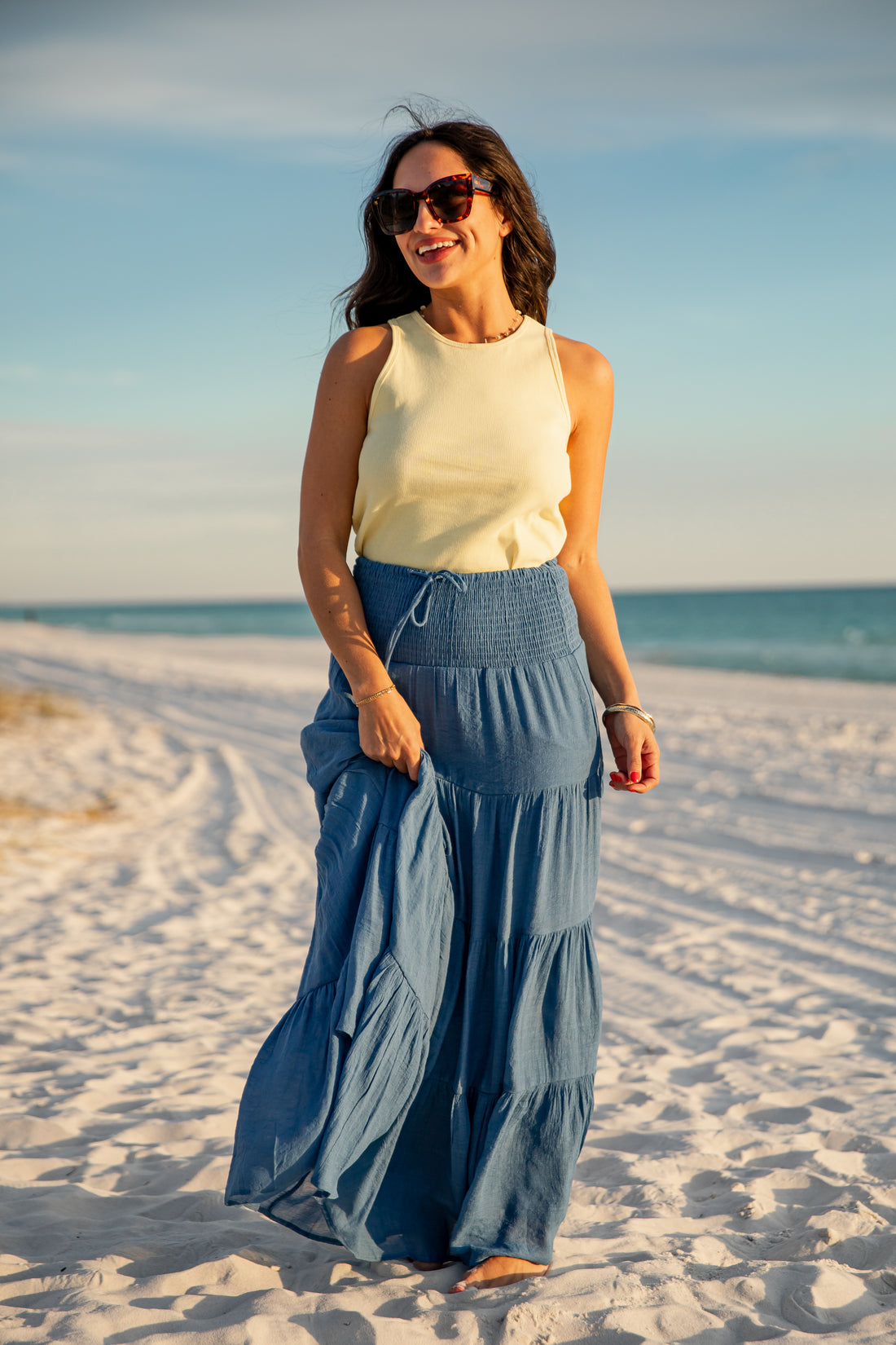 Woman in a yellow top and blue skirt standing on a sandy beach with ocean view.