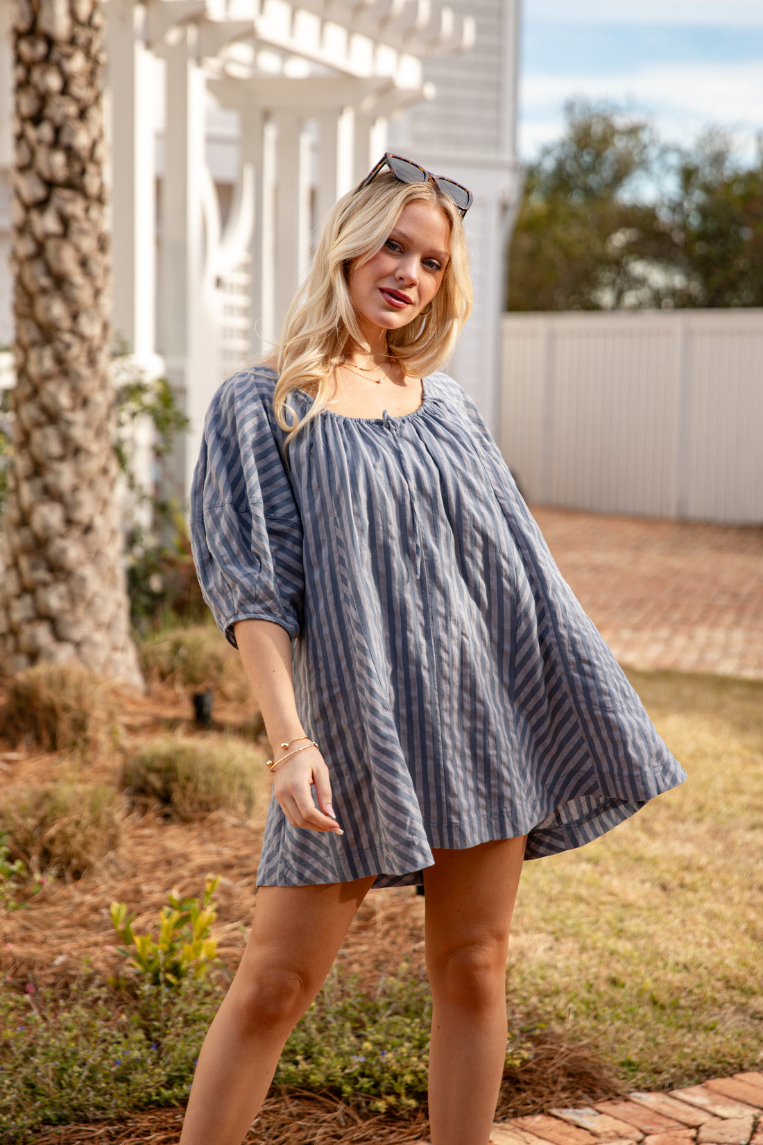Woman wearing a blue striped dress standing outdoors with trees and a white structure in the background.
