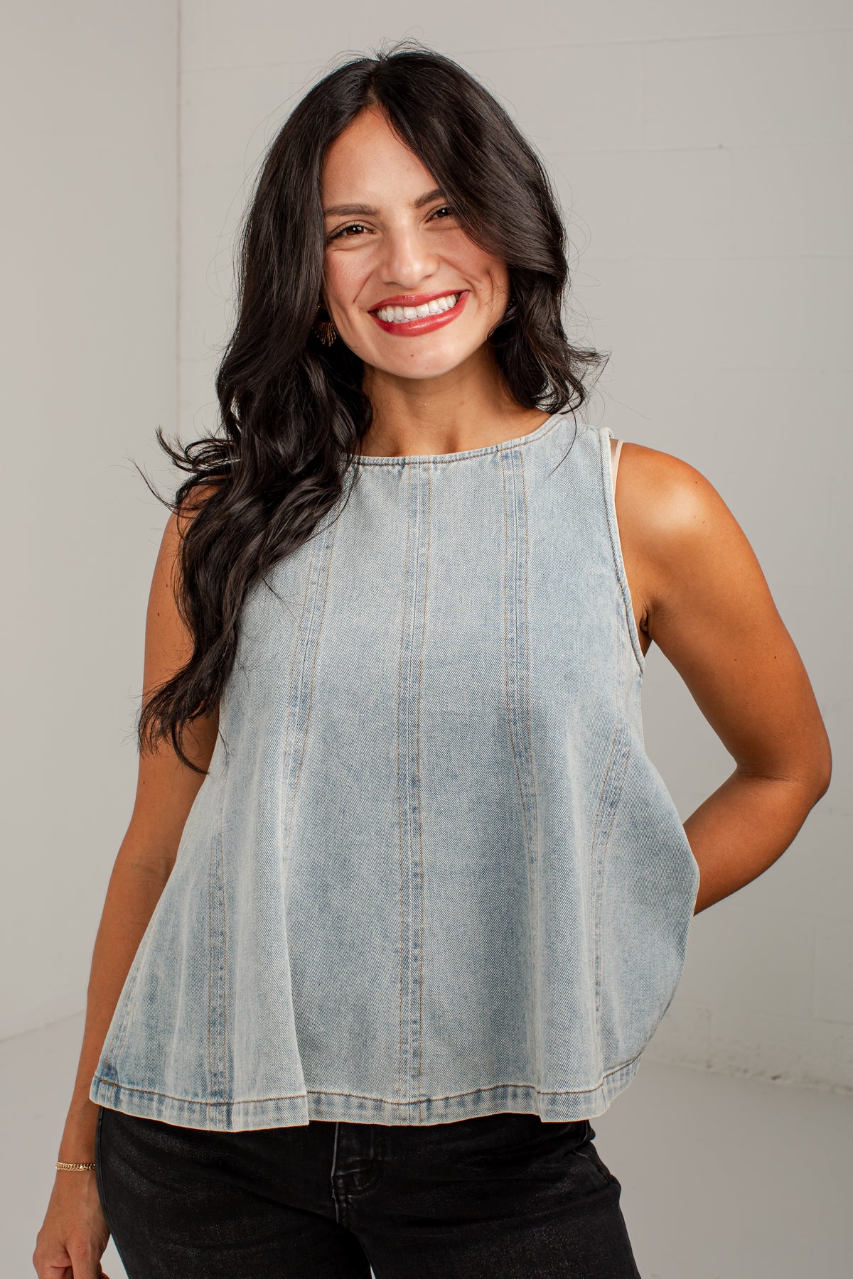 A smiling woman with long dark hair wearing a light-wash sleeveless denim top and black jeans, standing against a plain light gray wall.