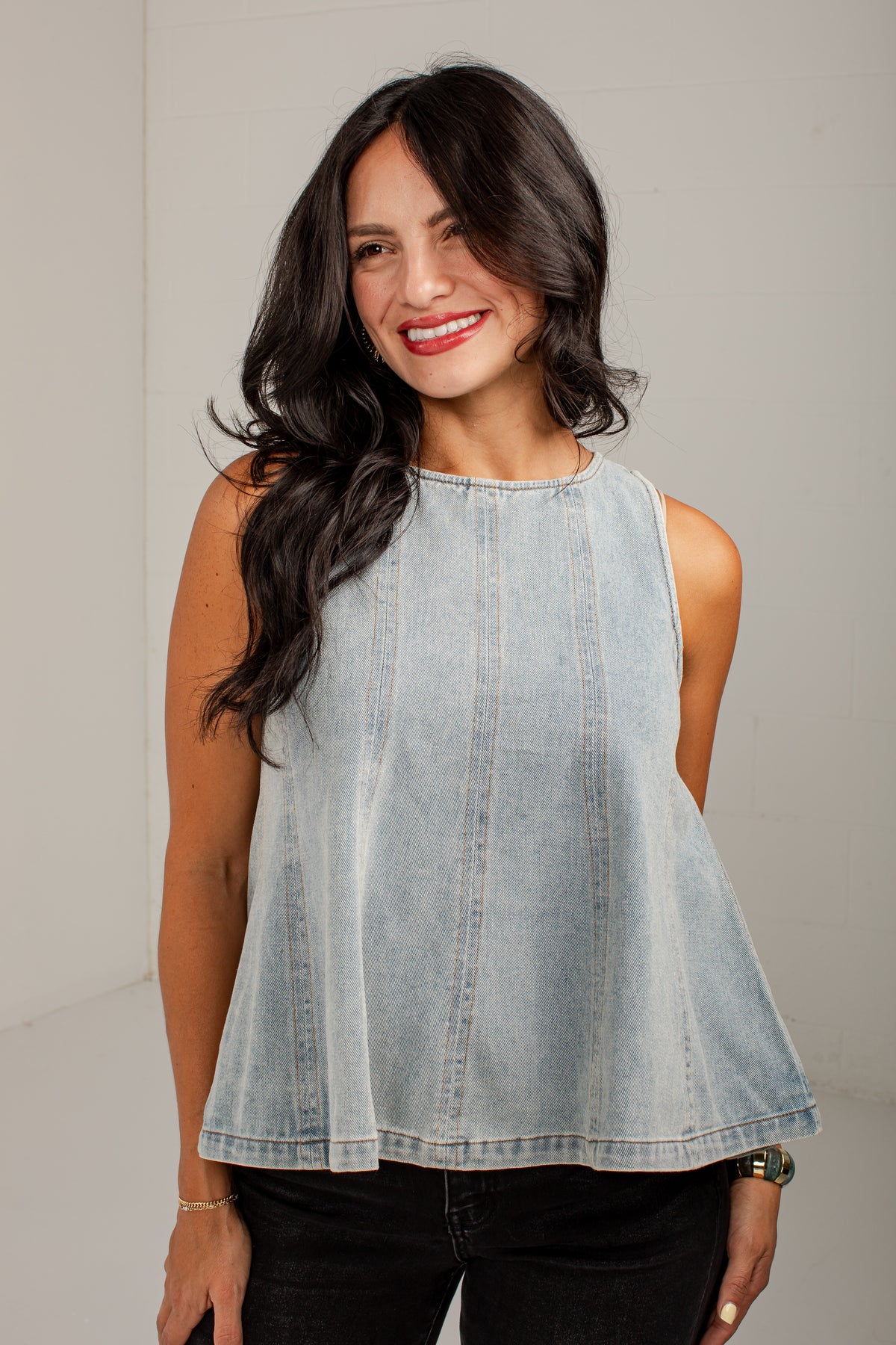 A woman modeling a sleeveless light-wash denim top with a flared peplum silhouette, paired with black pants, against a neutral studio background.