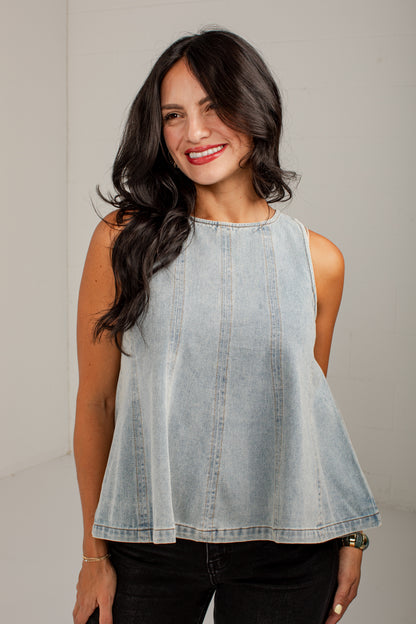 A woman modeling a sleeveless light-wash denim top with a flared peplum silhouette, paired with black pants, against a neutral studio background.