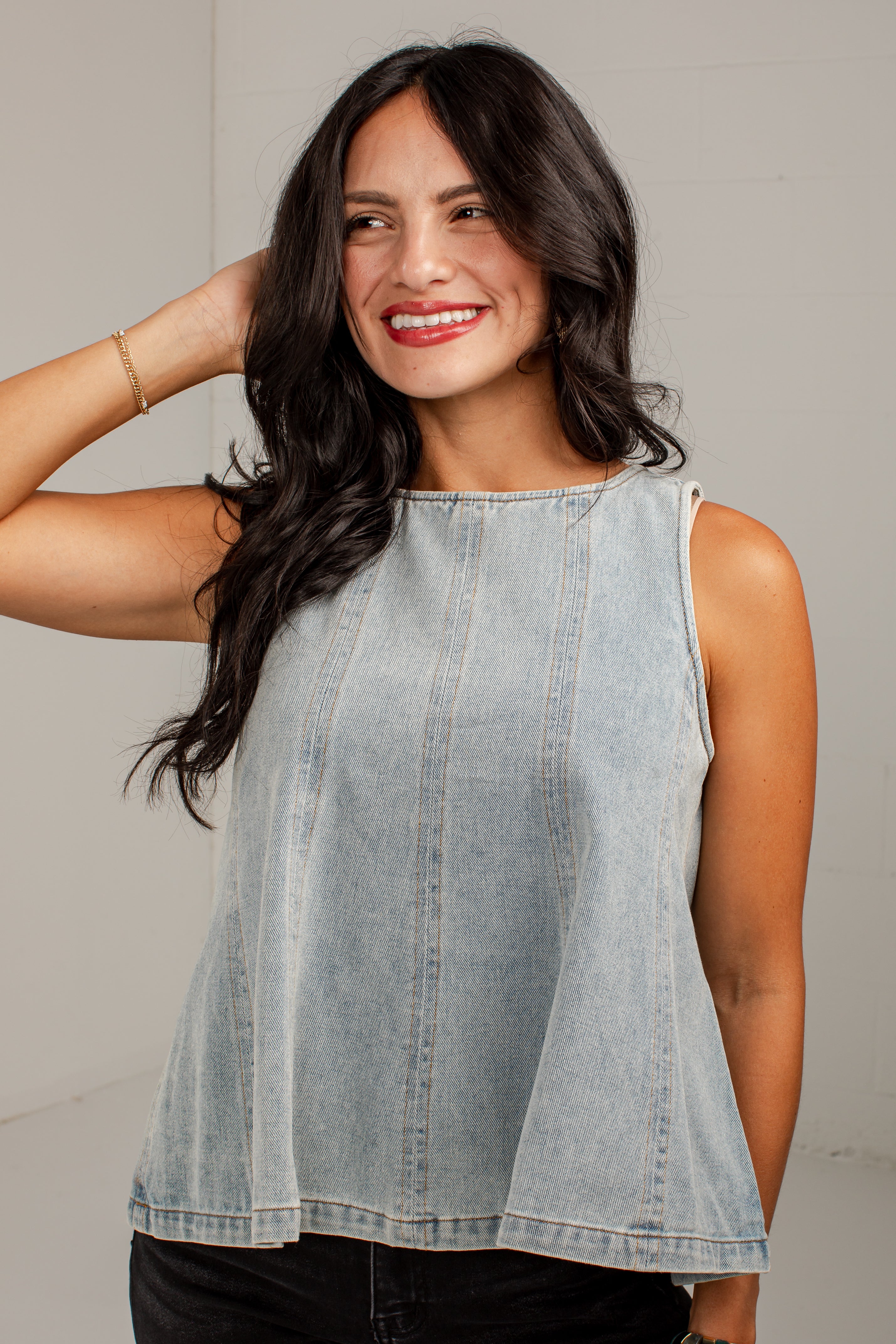 A woman with long dark hair wearing a sleeveless, light-wash denim peplum top, posing against a neutral light gray background.