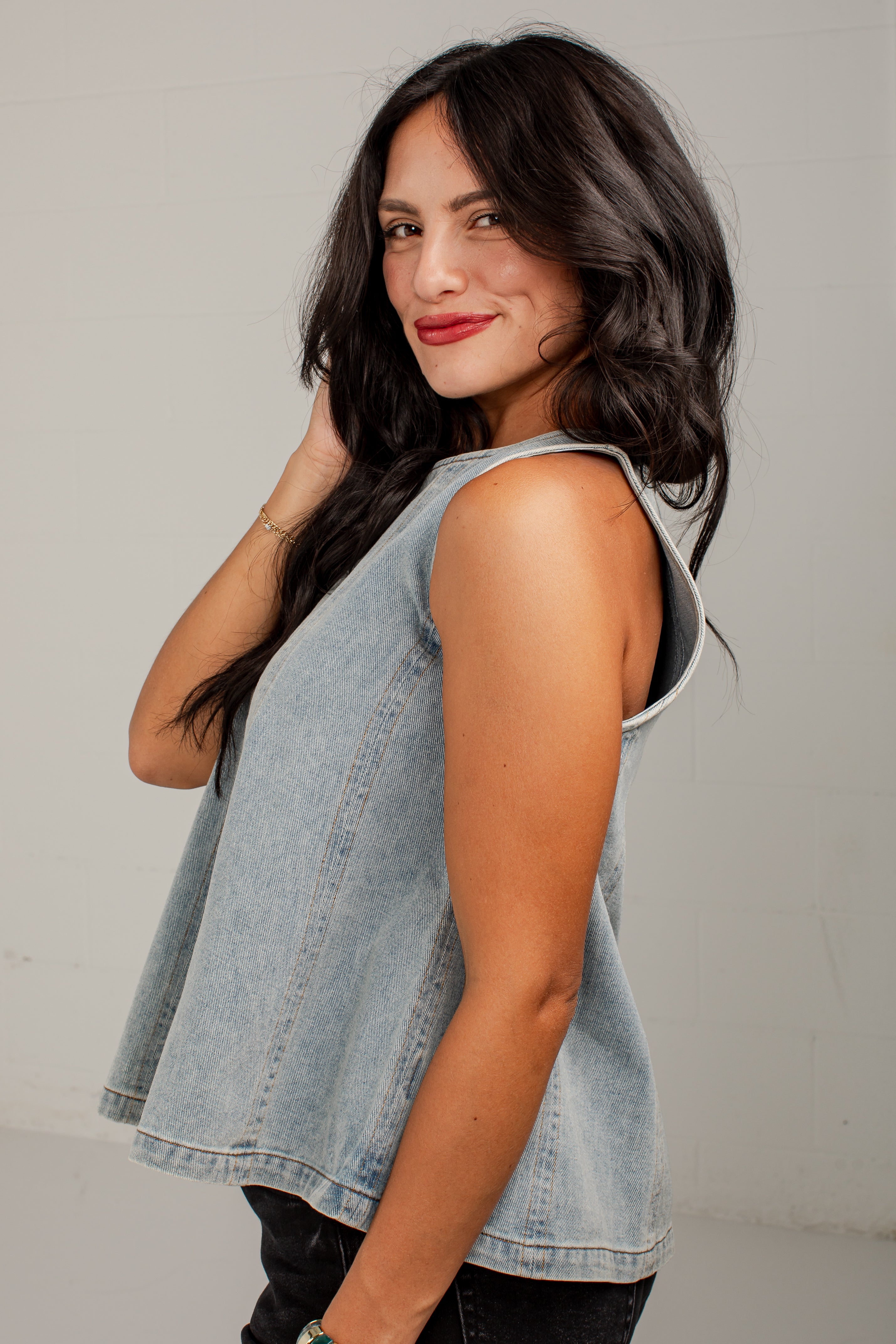 A woman wearing a sleeveless light-wash denim vest, photographed from the side and back against a plain light gray wall.