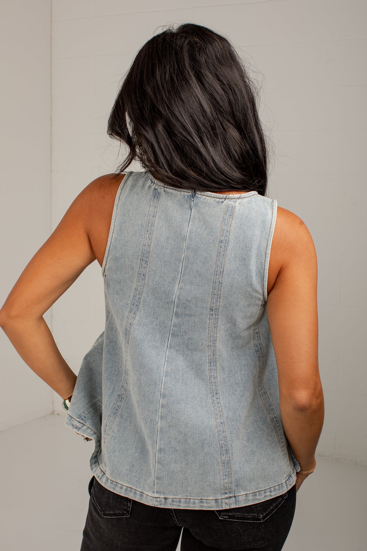 A woman viewed from behind wearing a sleeveless light-wash denim top with vertical seam detailing and dark jeans, set against a plain light gray background.