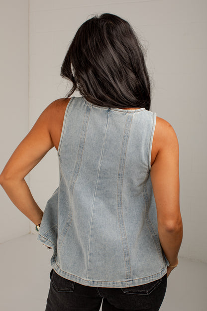 A woman viewed from behind wearing a sleeveless light-wash denim top with vertical seam detailing and dark jeans, set against a plain light gray background.