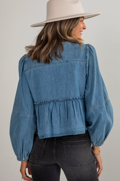 Woman wearing a blue denim blouse with ruffled details and a beige hat on a plain background