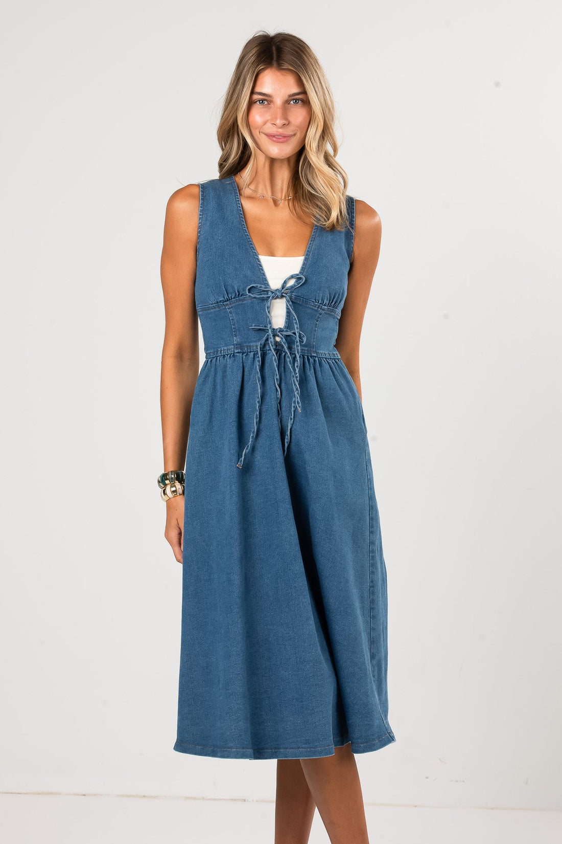 A woman modeling a sleeveless blue denim midi dress with a tie-front detail over a white undergarment, standing against a plain white studio background.