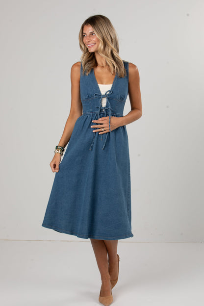 A woman modeling a sleeveless blue midi dress with a tie-front detail and a white underlayer visible at the neckline, standing against a plain light gray studio background.