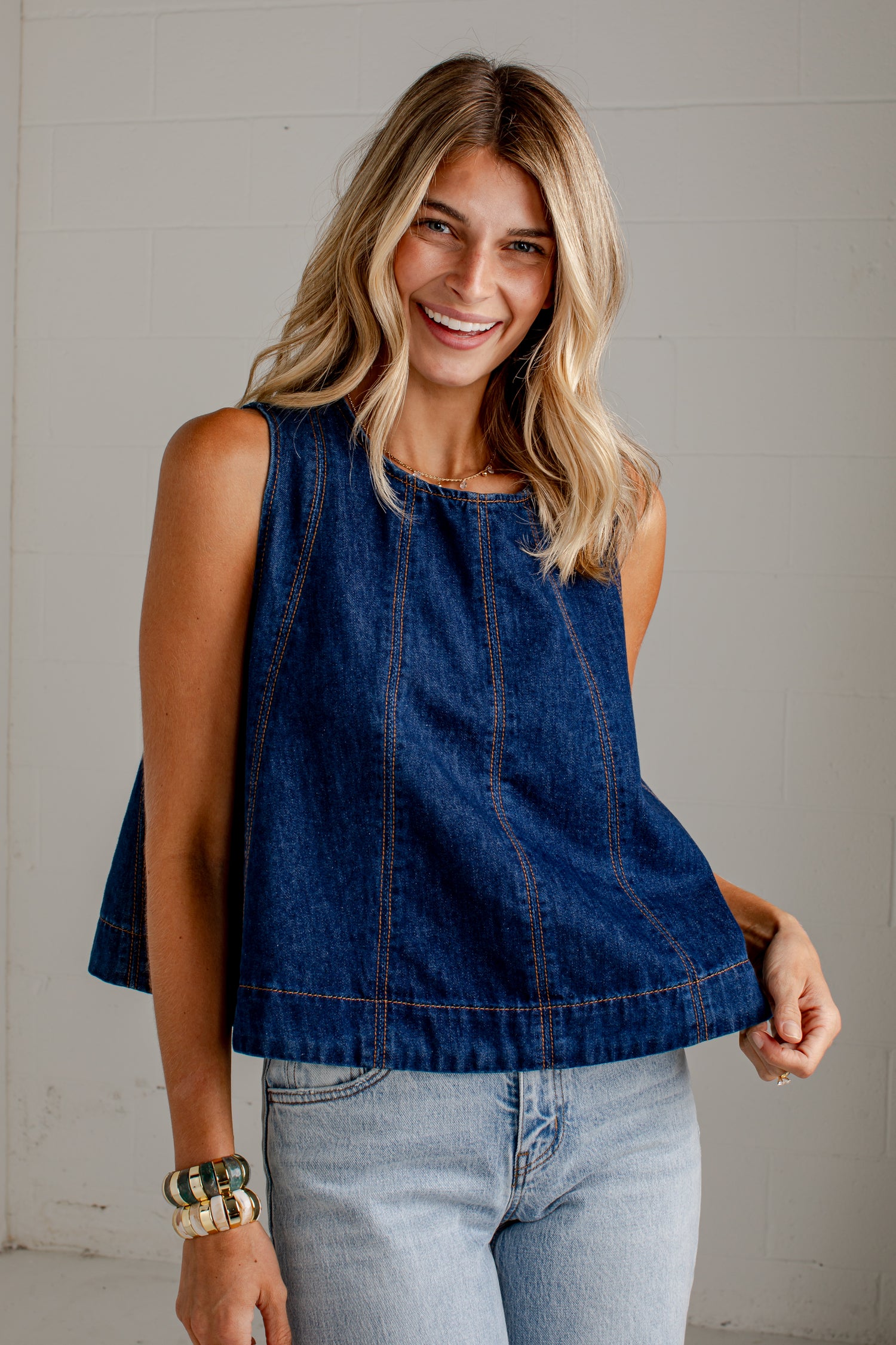 A woman wearing a sleeveless dark blue denim top with light-wash jeans, standing against a light gray wall.
