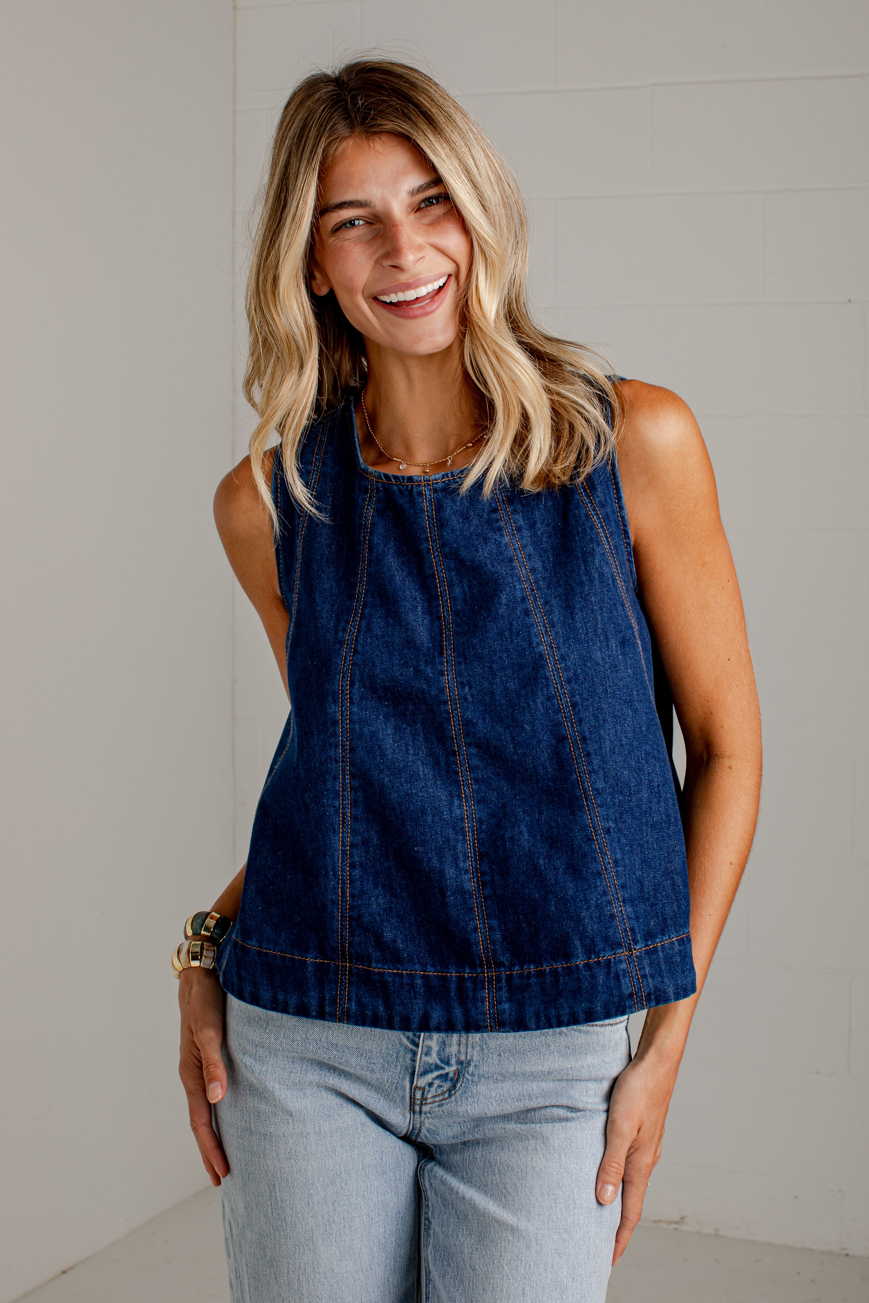 A woman wearing a sleeveless dark blue denim top and light-wash jeans, standing against a light gray wall.