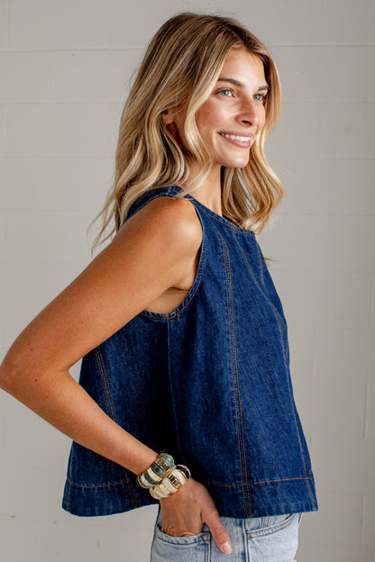 A woman wearing a sleeveless dark blue denim top, paired with light-wash jeans and stacked bracelets, posing against a light gray paneled wall.