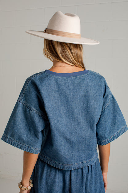 A woman shown from behind wearing a blue denim short-sleeve top and matching skirt, paired with a wide-brimmed cream hat, against a light gray brick wall.