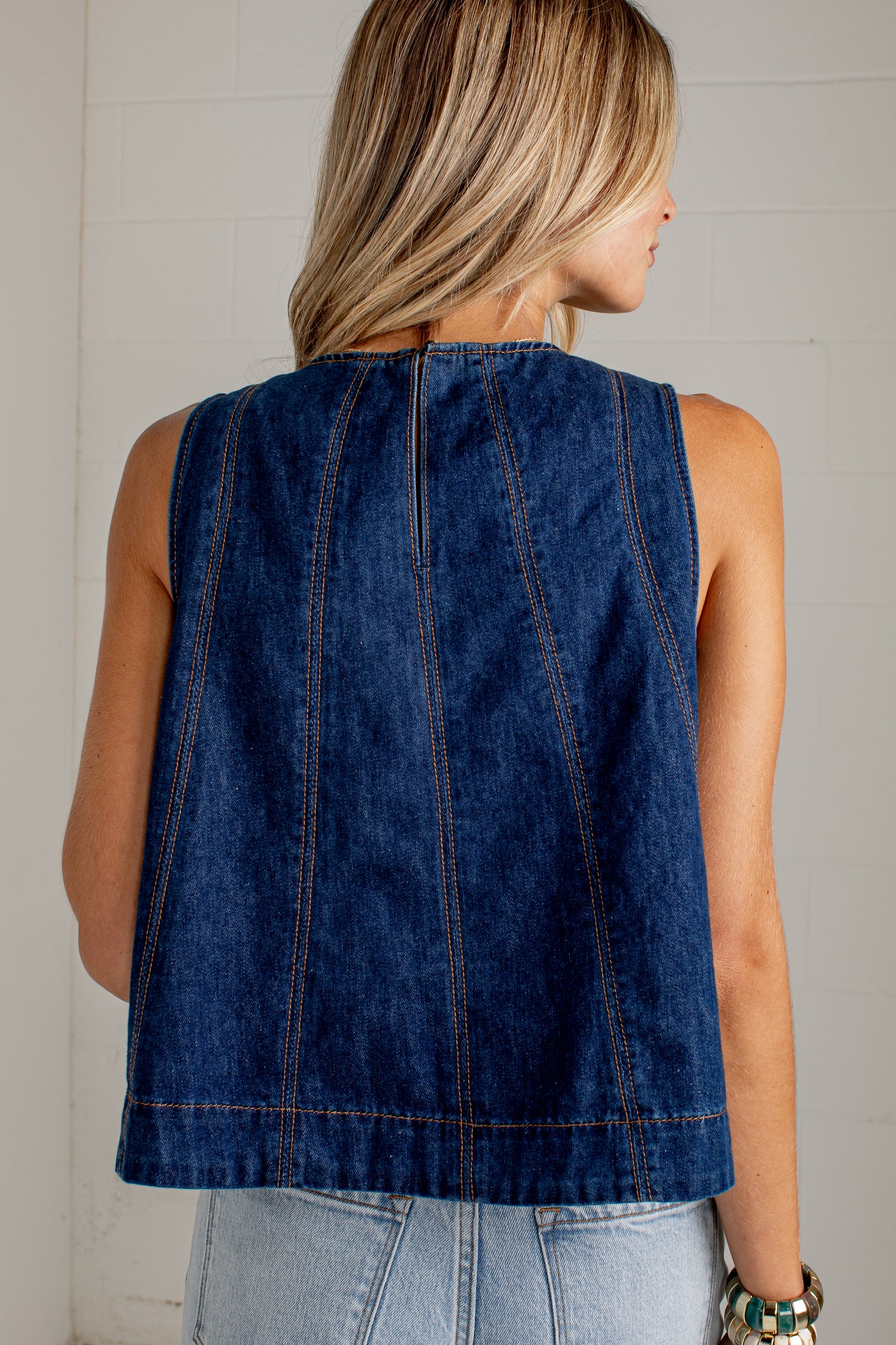 A woman shown from the back wearing a sleeveless dark blue denim vest with vertical seam detailing, paired with light blue jeans against a white brick wall.