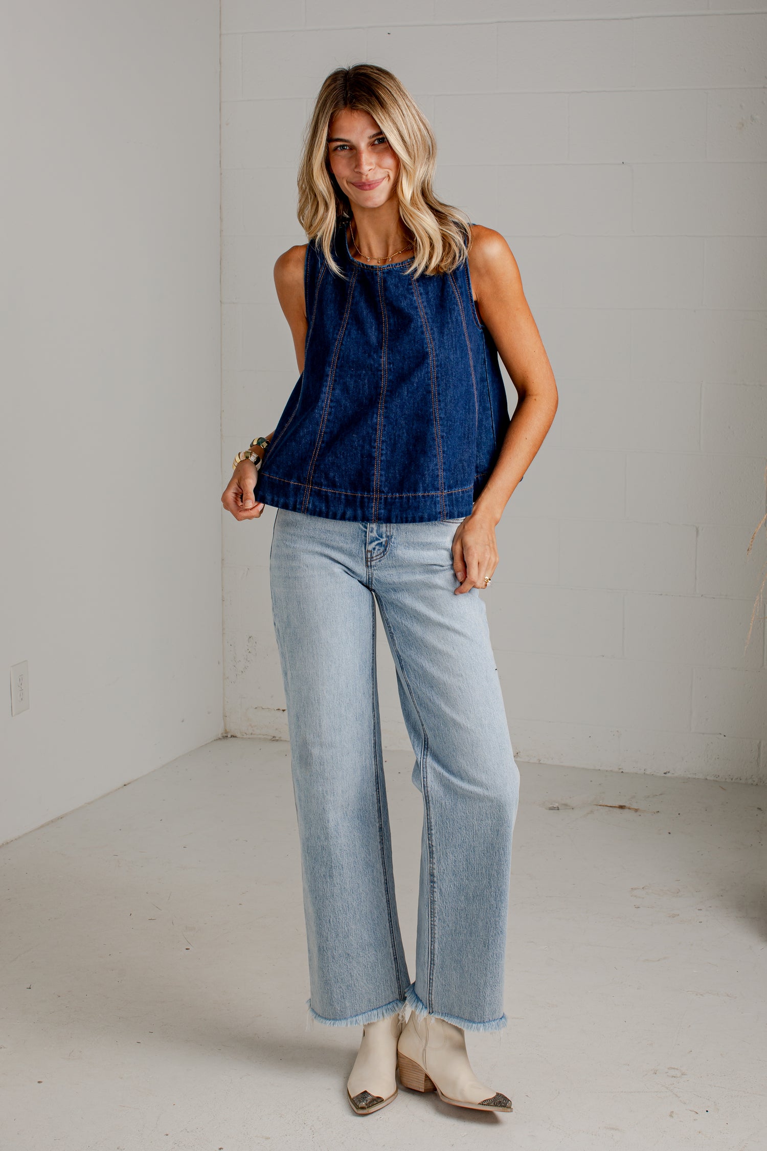 A woman modeling a sleeveless dark blue denim top paired with light-wash wide-leg jeans and beige heeled sandals, standing in a bright white studio space.