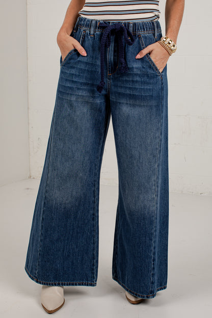 A person wearing dark blue wide-leg denim jeans with a drawstring waistband, standing against a white wall.