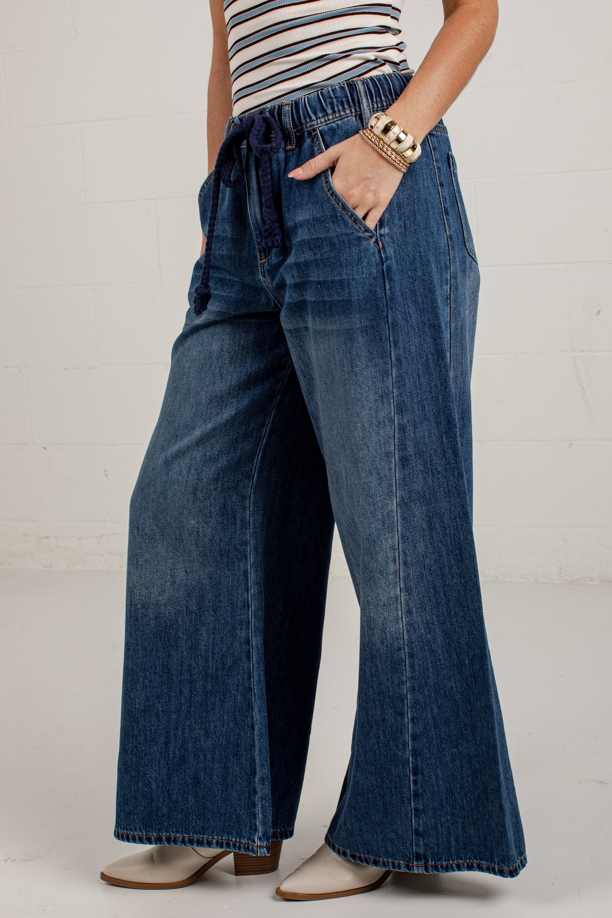 A person wearing blue wide-leg denim jeans with a drawstring waistband, paired with a striped top and beige heeled shoes, standing against a white brick wall.