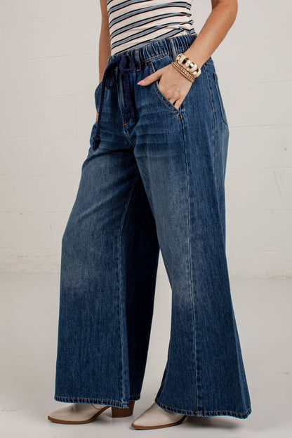 A person wearing blue wide-leg denim jeans with a drawstring waistband, paired with a striped top and beige heeled shoes, standing against a white brick wall.