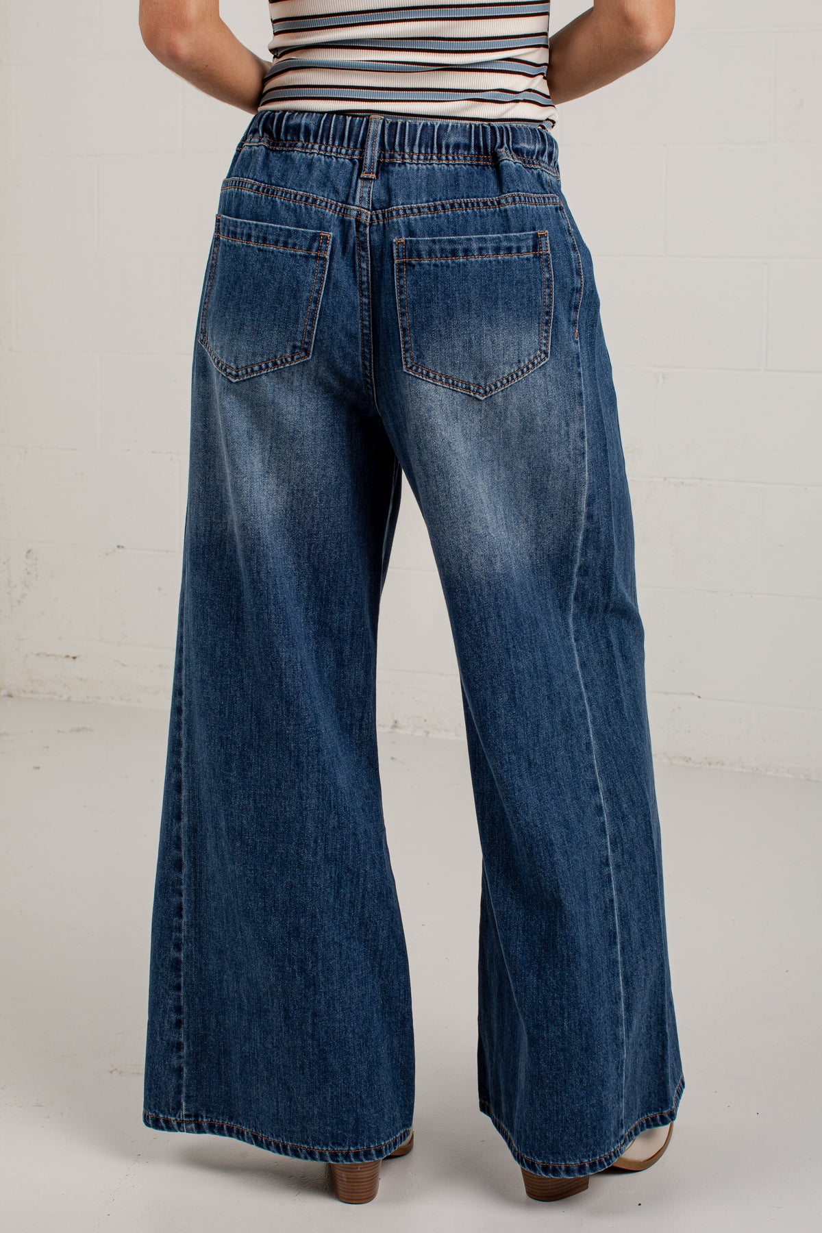 Back view of a person wearing high-waisted, wide-leg blue denim jeans with a striped top, standing against a white brick wall.