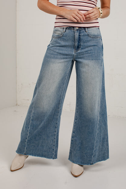 A person wearing light-to-medium blue wide-leg denim jeans, a red-and-white striped top, and white pointed-toe heels, standing against a plain white wall.