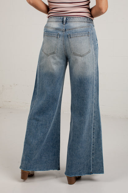 A person shown from the back wearing wide-leg blue denim jeans with a faded wash, paired with a striped top and brown heels, against a light gray studio background.