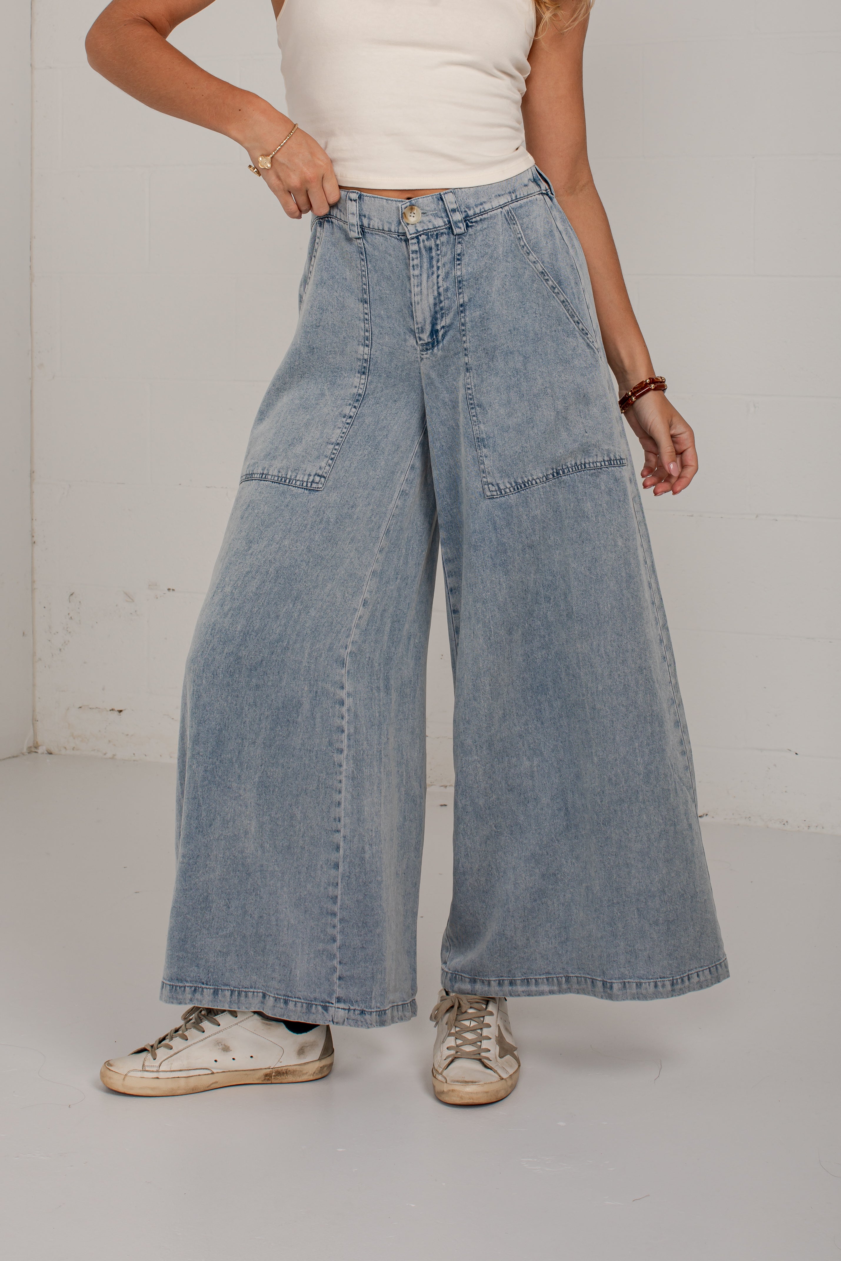 A person wearing light-wash wide-leg denim jeans with a cream ribbed tank top and distressed white sneakers, standing against a white wall.