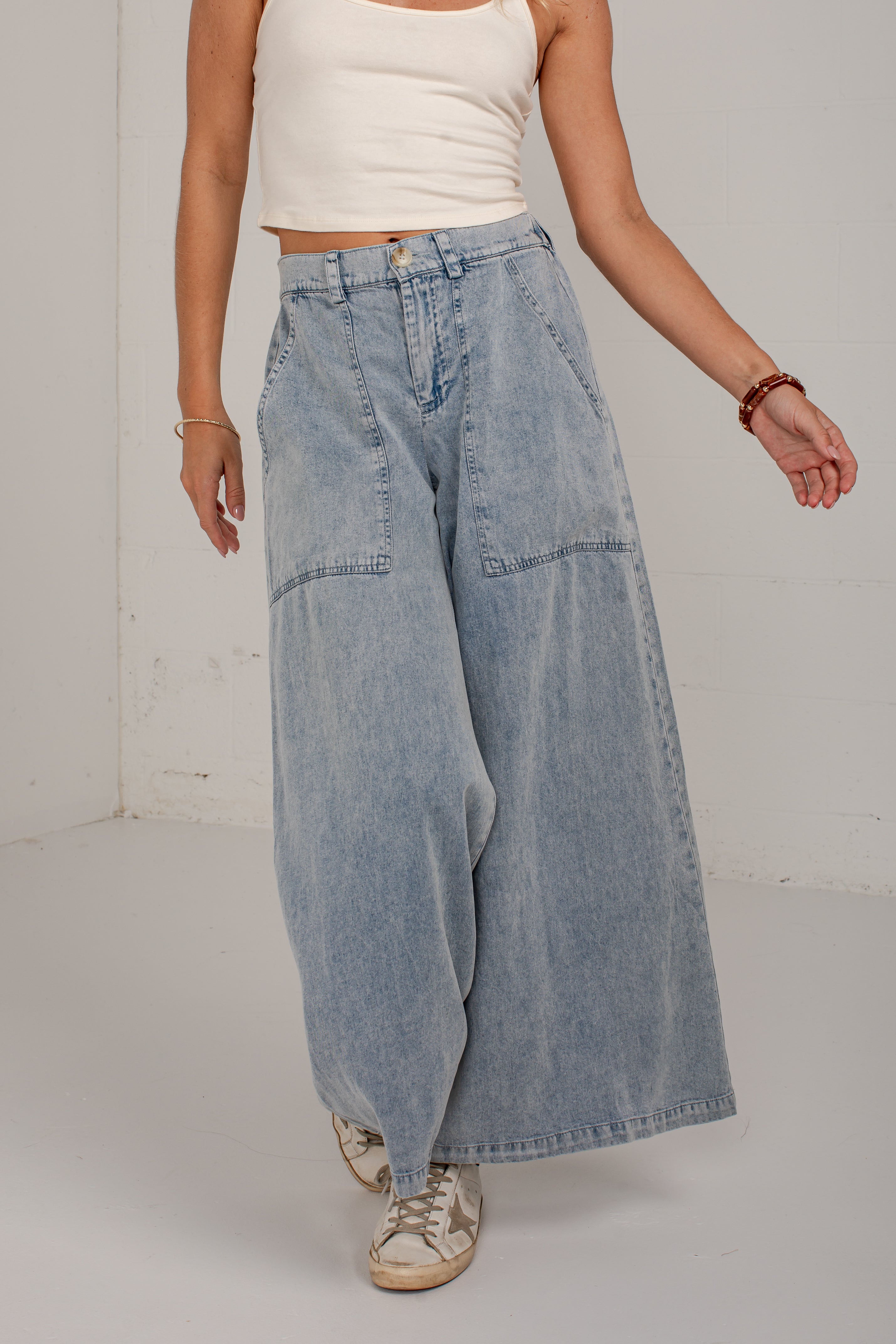 A model wearing light-wash, wide-leg denim pants with front patch pockets, styled with a white ribbed tank top and distressed sneakers.