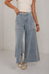 A model wearing light-wash blue wide-leg denim pants with a cream ribbed tank top and distressed sneakers, standing against a white wall.