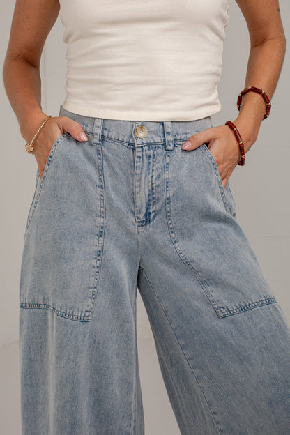A close-up of a person wearing light blue wide-leg denim jeans with a white ribbed tank top, standing against a neutral gray background.