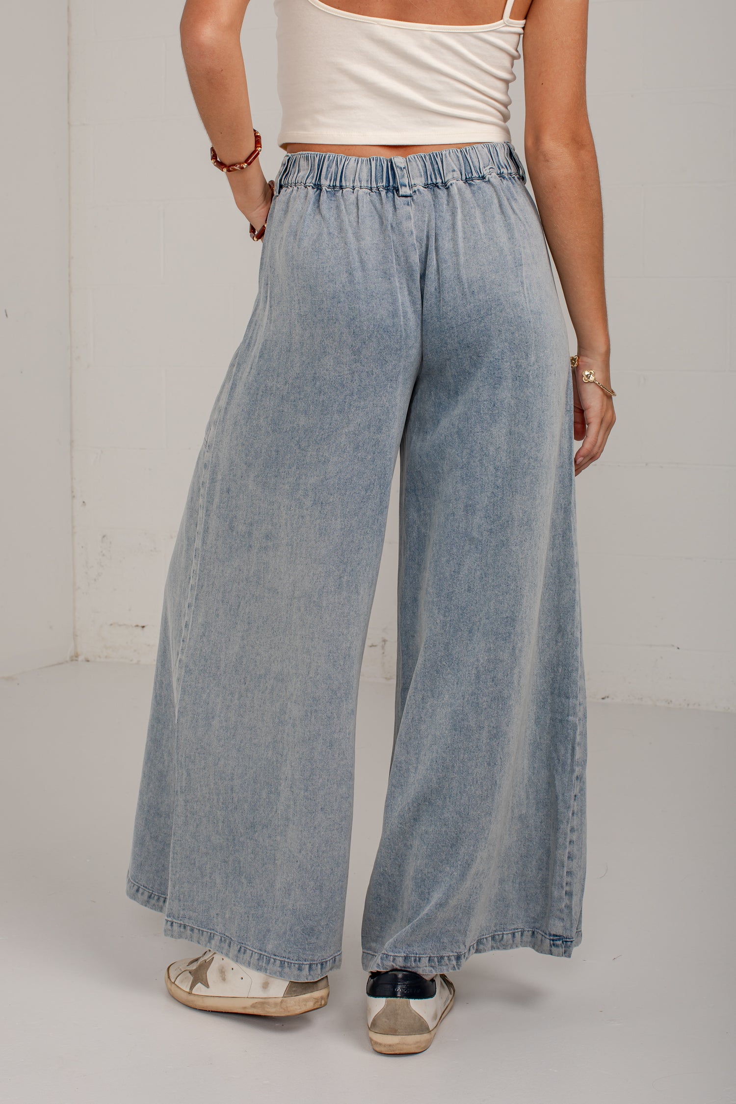 A person wearing light-wash wide-leg denim pants with an elastic waistband, paired with a white cropped tank top and white sneakers, standing against a plain off-white wall.