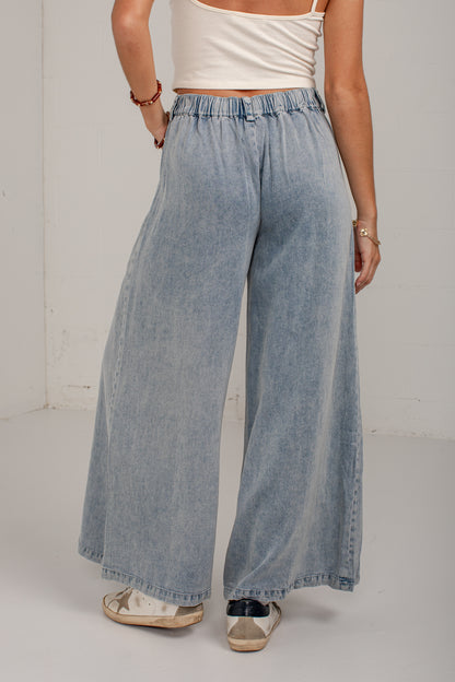 A person wearing light-wash wide-leg denim pants with an elastic waistband, paired with a white cropped tank top and white sneakers, standing against a plain off-white wall.