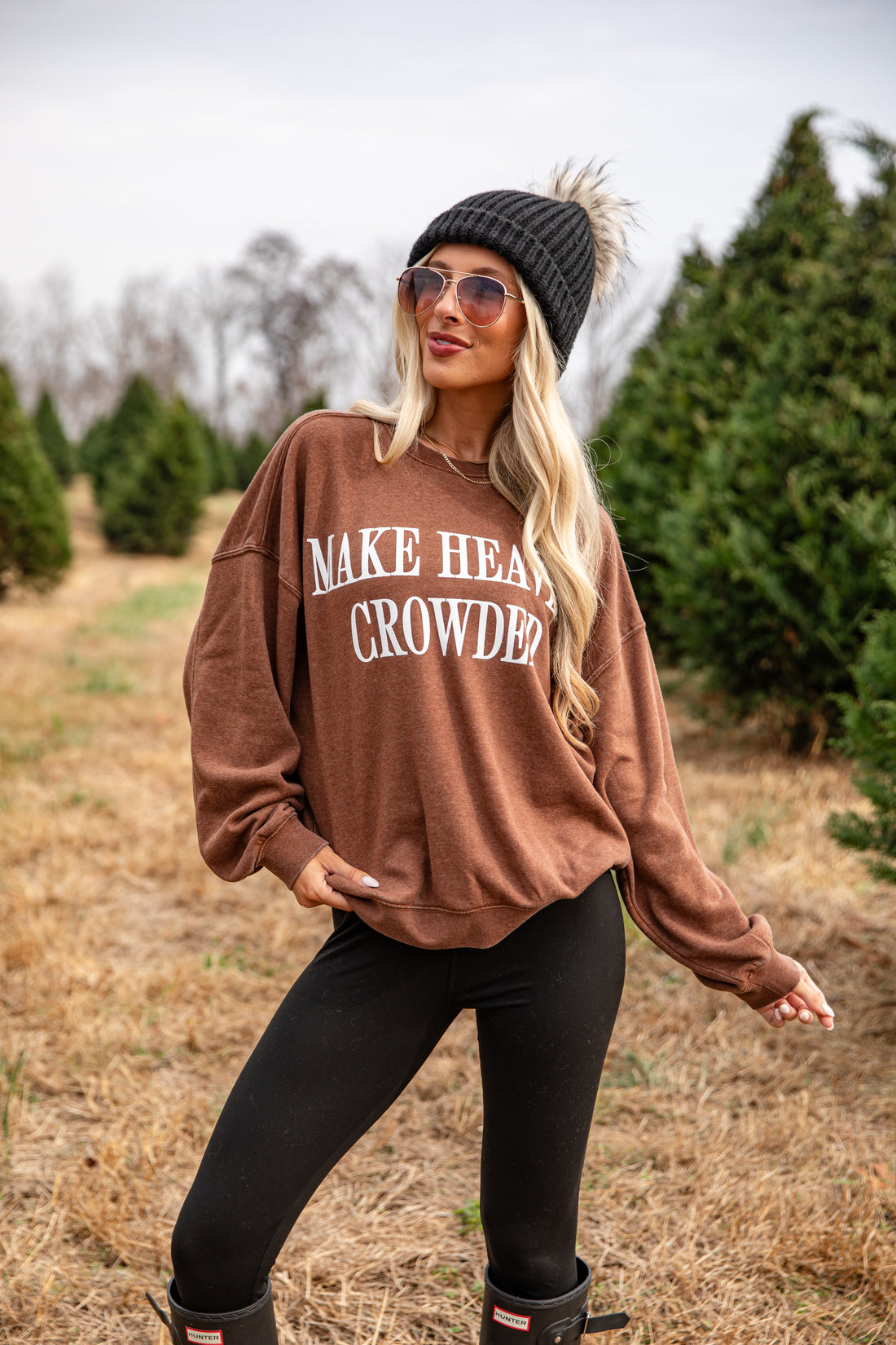 Woman wearing a brown sweatshirt with text, sunglasses, and a beanie in a field.