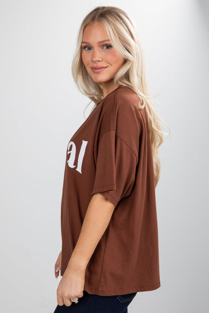Woman wearing a brown oversized t-shirt with white text on a plain background