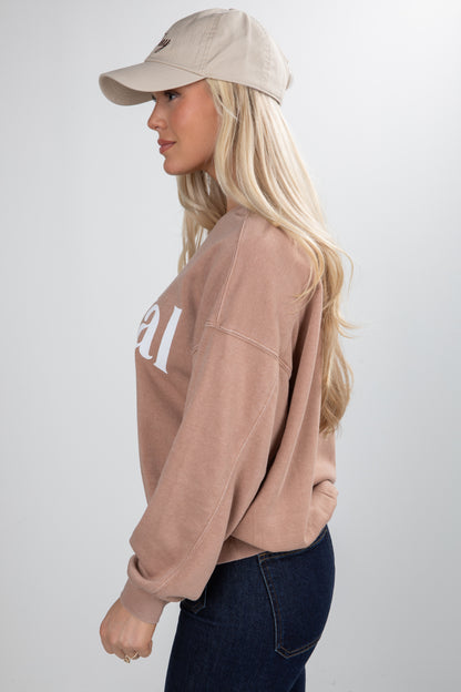 Woman wearing a beige sweatshirt with text, dark jeans, and a beige cap on a light gray background