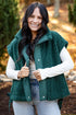 A woman wearing a dark green sherpa fleece vest over a white long-sleeve shirt and light blue jeans, standing outdoors in a park-like setting with trees and autumn foliage in the background.