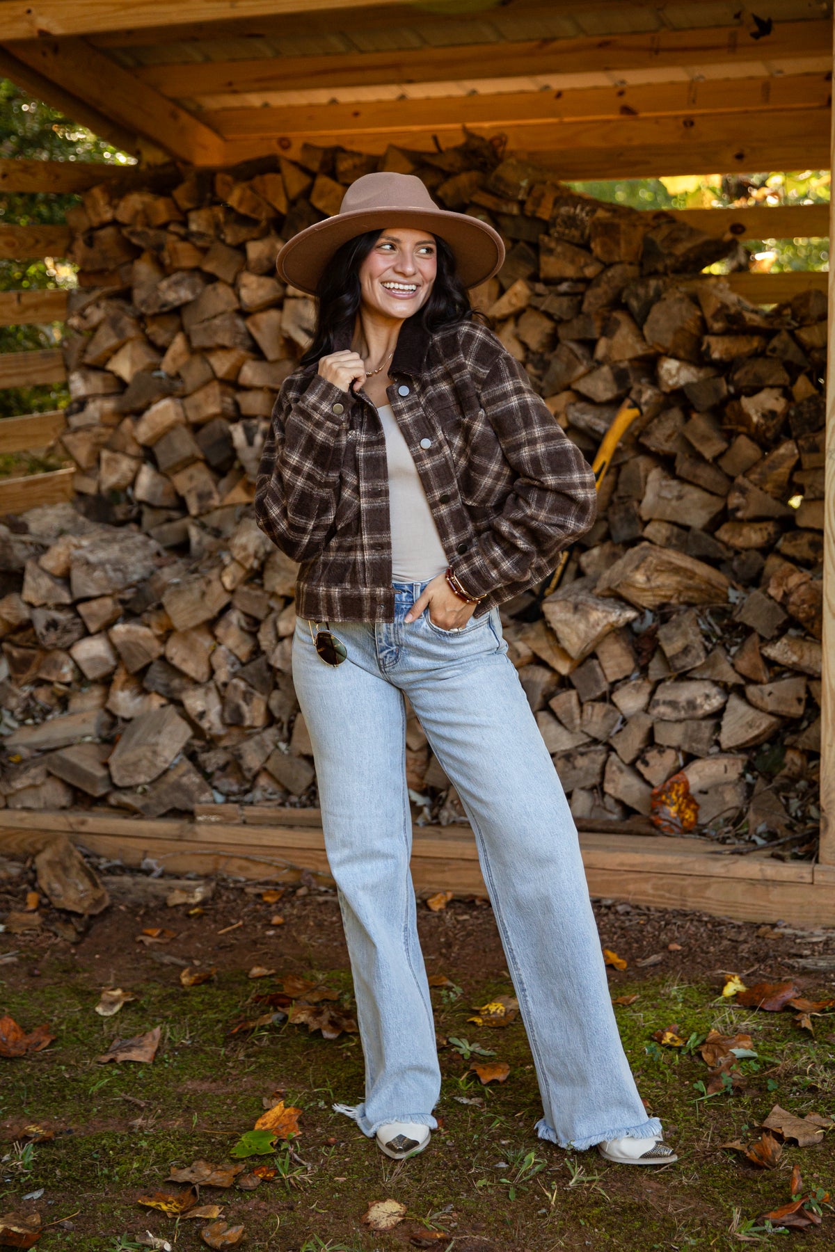 A woman stands outdoors in front of a stacked woodpile under a wooden shelter, wearing a brown plaid flannel shirt, a wide-brim tan hat, a light beige top, and light-wash blue jeans.