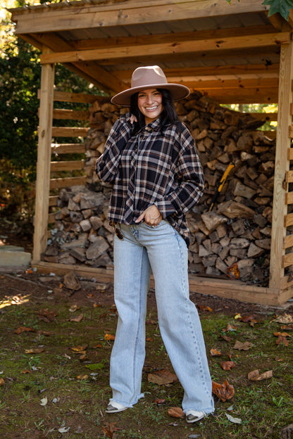 A woman stands outdoors in front of a wooden firewood shelter, wearing a brown wide-brim hat, a black and brown plaid button-up shirt, and light-wash blue jeans.