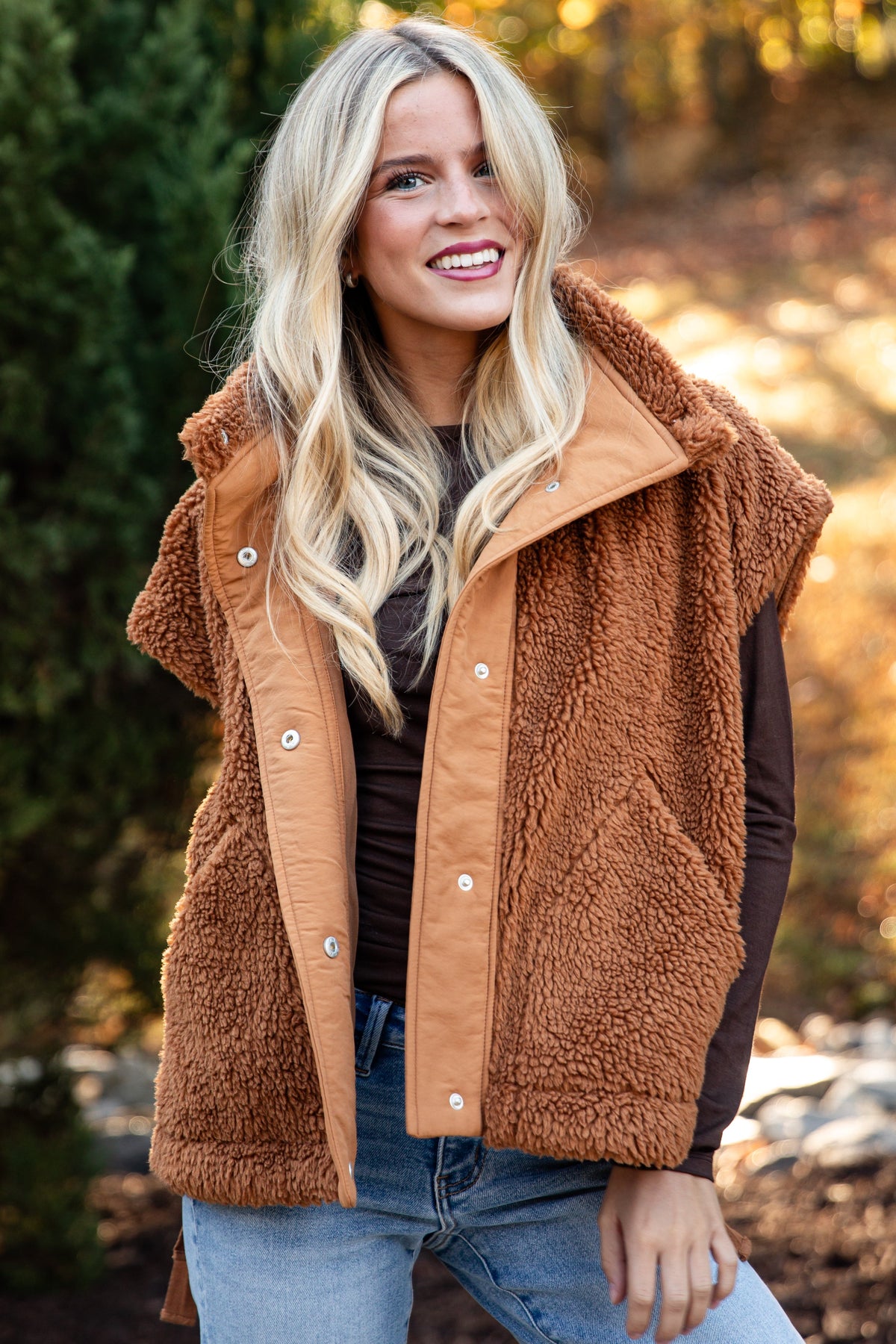 A smiling blonde woman wearing a brown sherpa fleece vest over a dark top and blue jeans, standing outdoors in a wooded setting with trees and sunlight in the background.