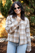 A woman wearing a beige and brown plaid zip-up jacket with a dark brown collar, styled with light blue jeans and sunglasses, standing outdoors in front of a blurred background of trees and foliage.