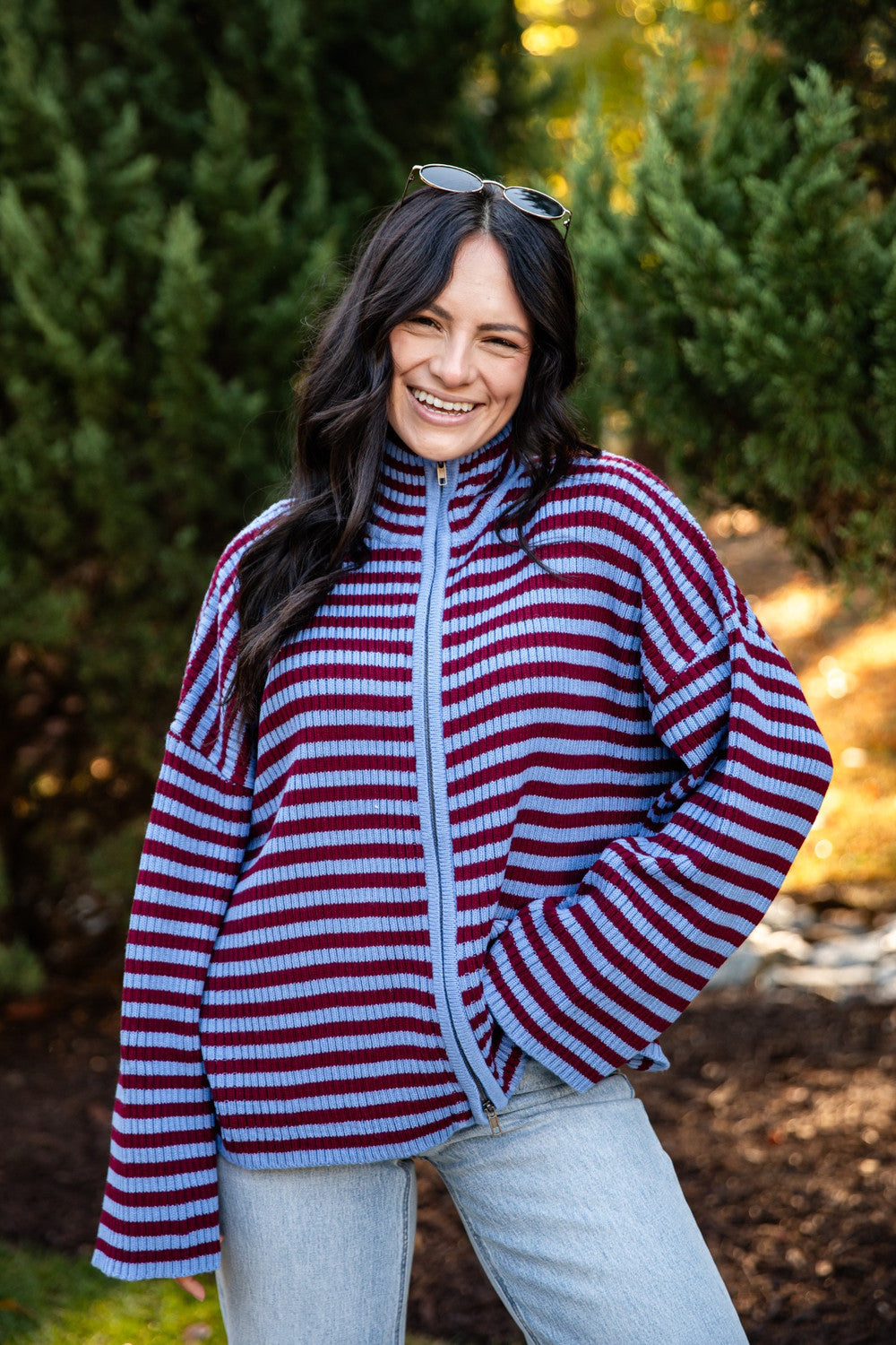 A woman smiling outdoors wearing a red and blue horizontally striped zip-up fleece jacket with a light blue zipper, paired with light-wash jeans.