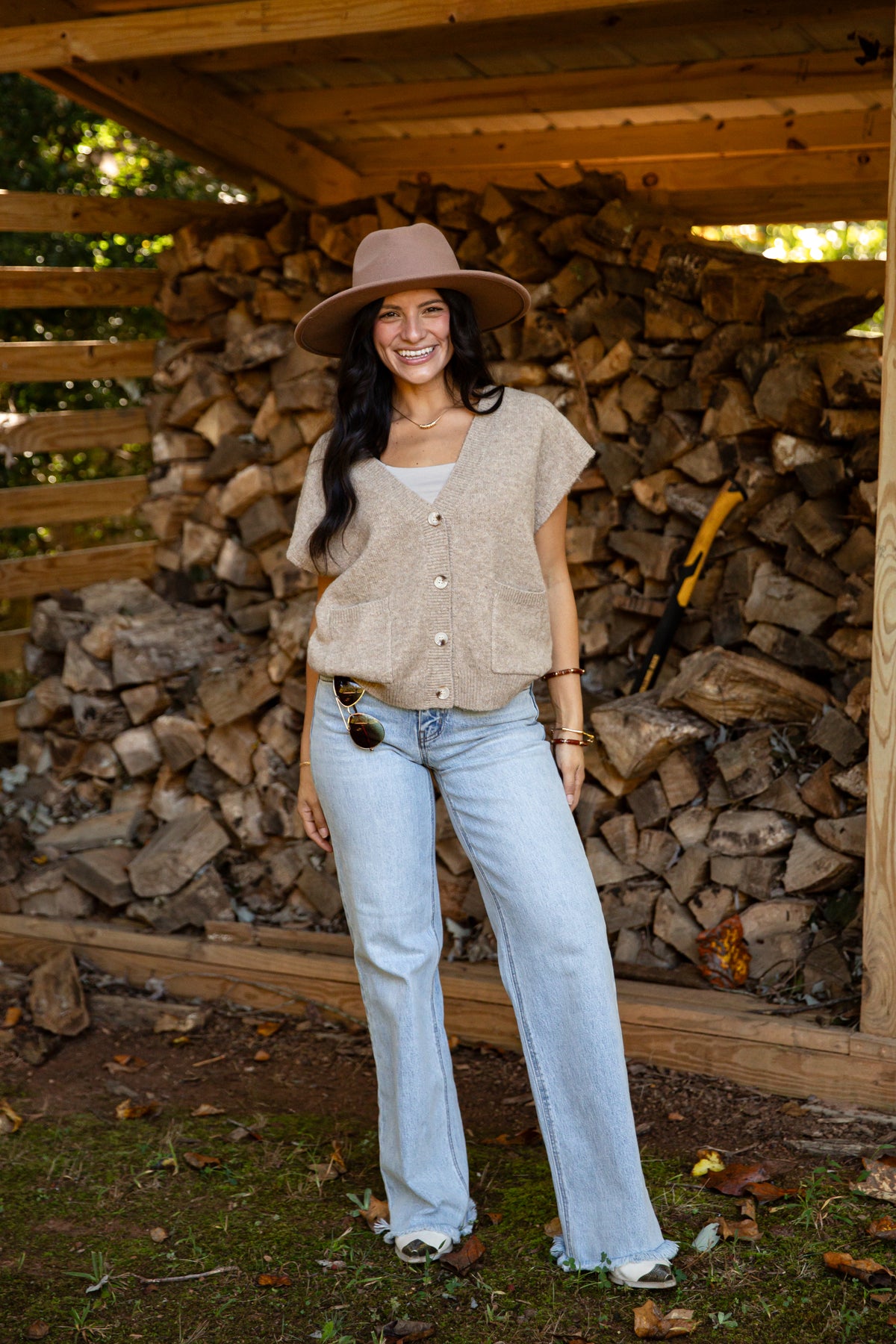 A woman stands outdoors in front of a stacked woodpile under a wooden shelter, wearing a wide-brim tan hat, a beige short-sleeve button-front top, light-wash blue jeans, and white sneakers.