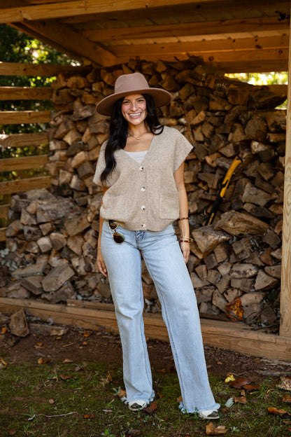 A woman stands outdoors in front of a stacked woodpile under a wooden shelter, wearing a wide-brim tan hat, a beige short-sleeve button-front top, light-wash blue jeans, and white sneakers.