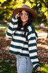 A woman wearing a dark green and white striped knit sweater, a brown wide-brim hat, and light-wash jeans, standing outdoors in a park with trees and fallen leaves in the background.
