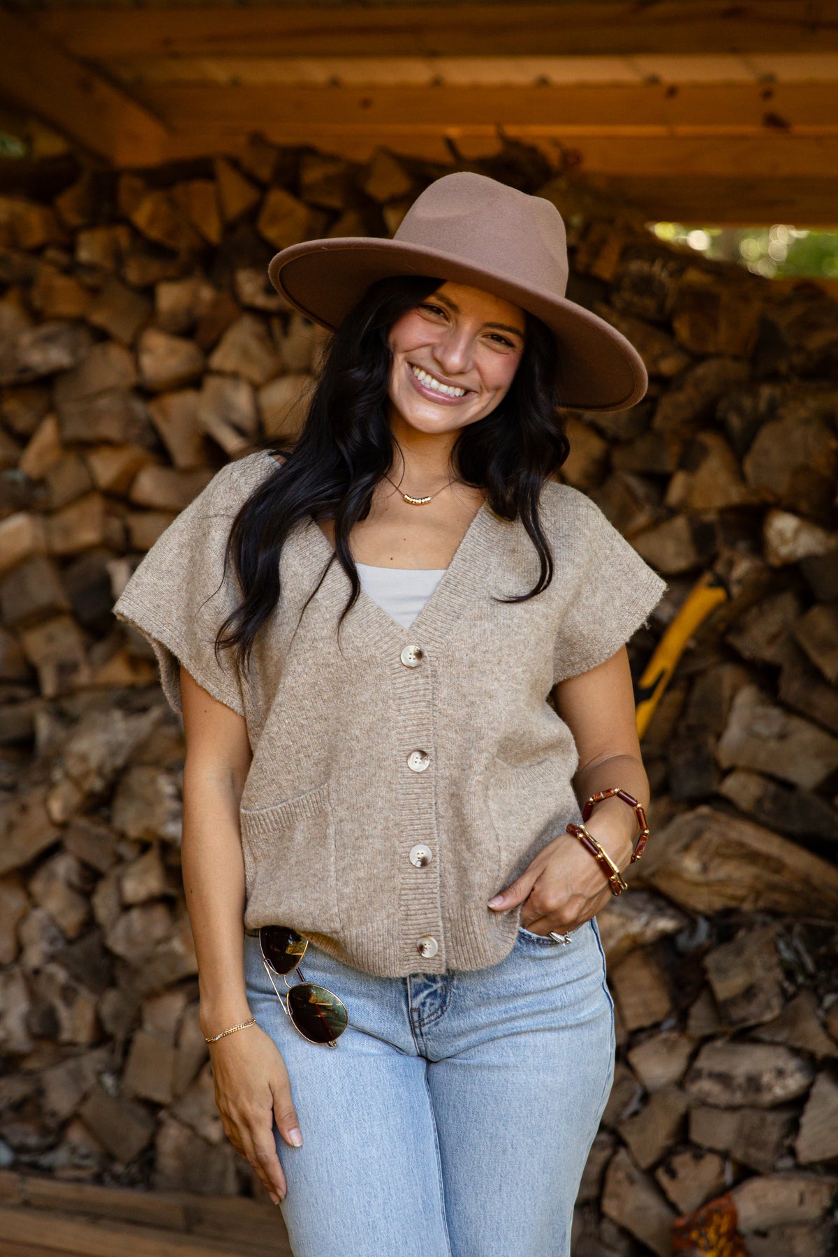 A woman smiling in front of a stacked woodpile, wearing a taupe wide-brim hat, a beige short-sleeve button-front knit top over a white tank top, and light-wash blue jeans.