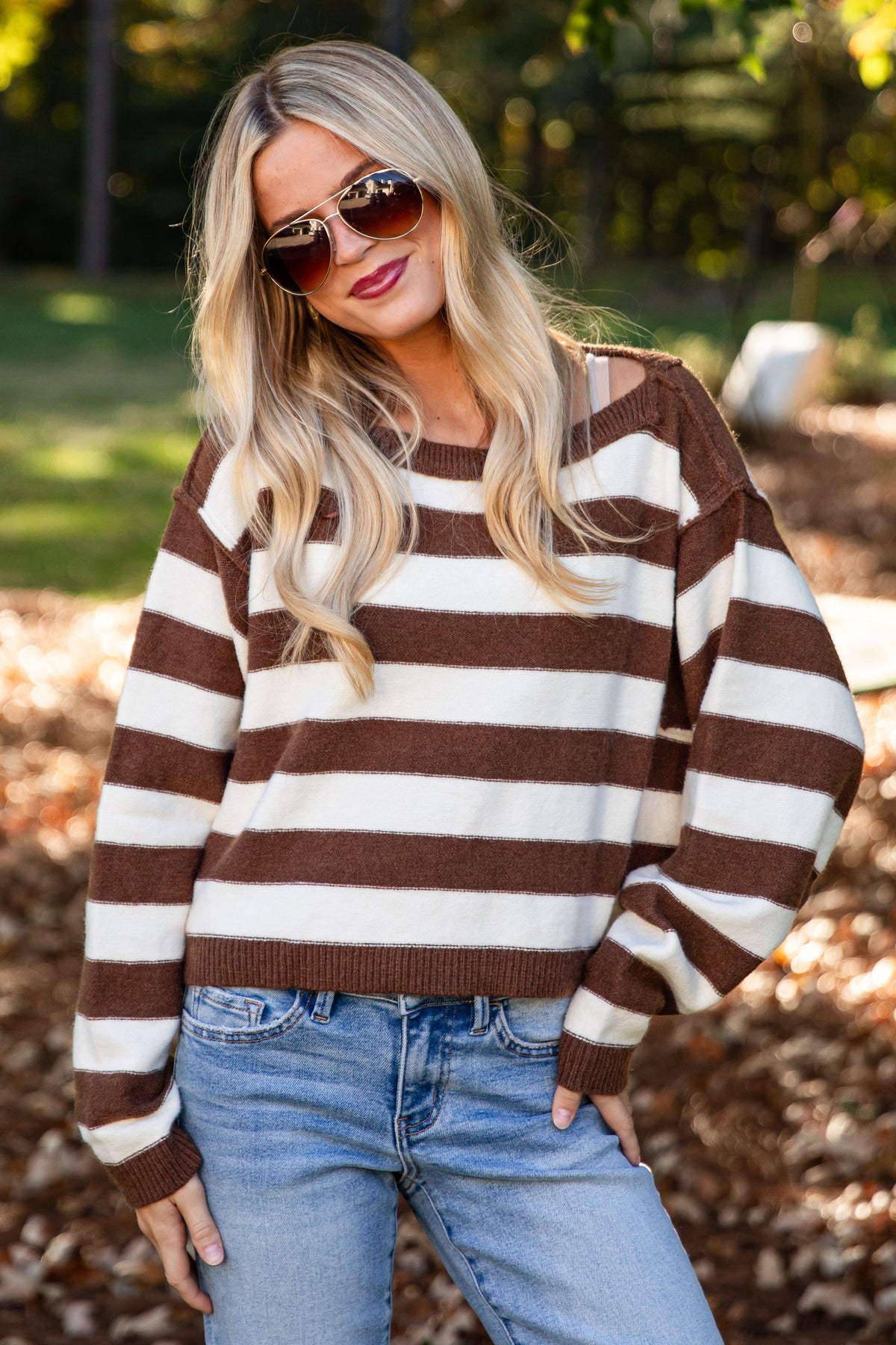 A woman wearing a brown and cream striped knit sweater and light blue jeans stands outdoors in a park with fallen leaves and trees in the background.