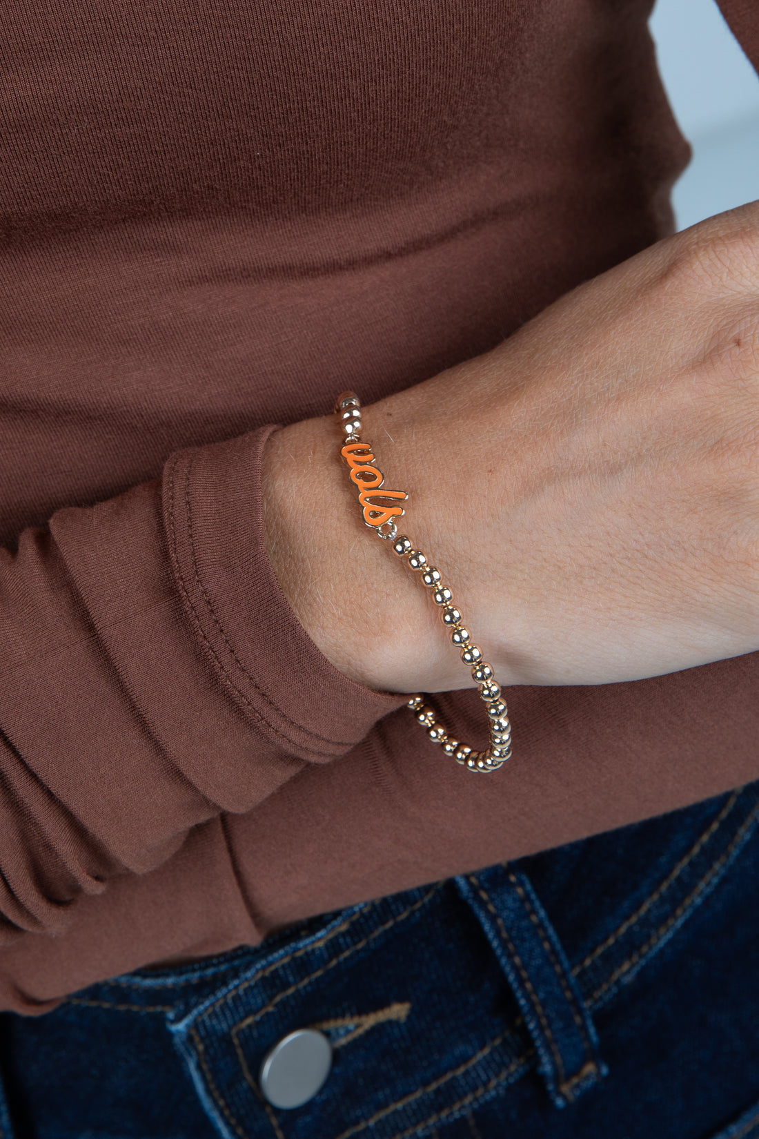 Close-up of a wrist wearing a bracelet with orange letters, against a brown shirt background.