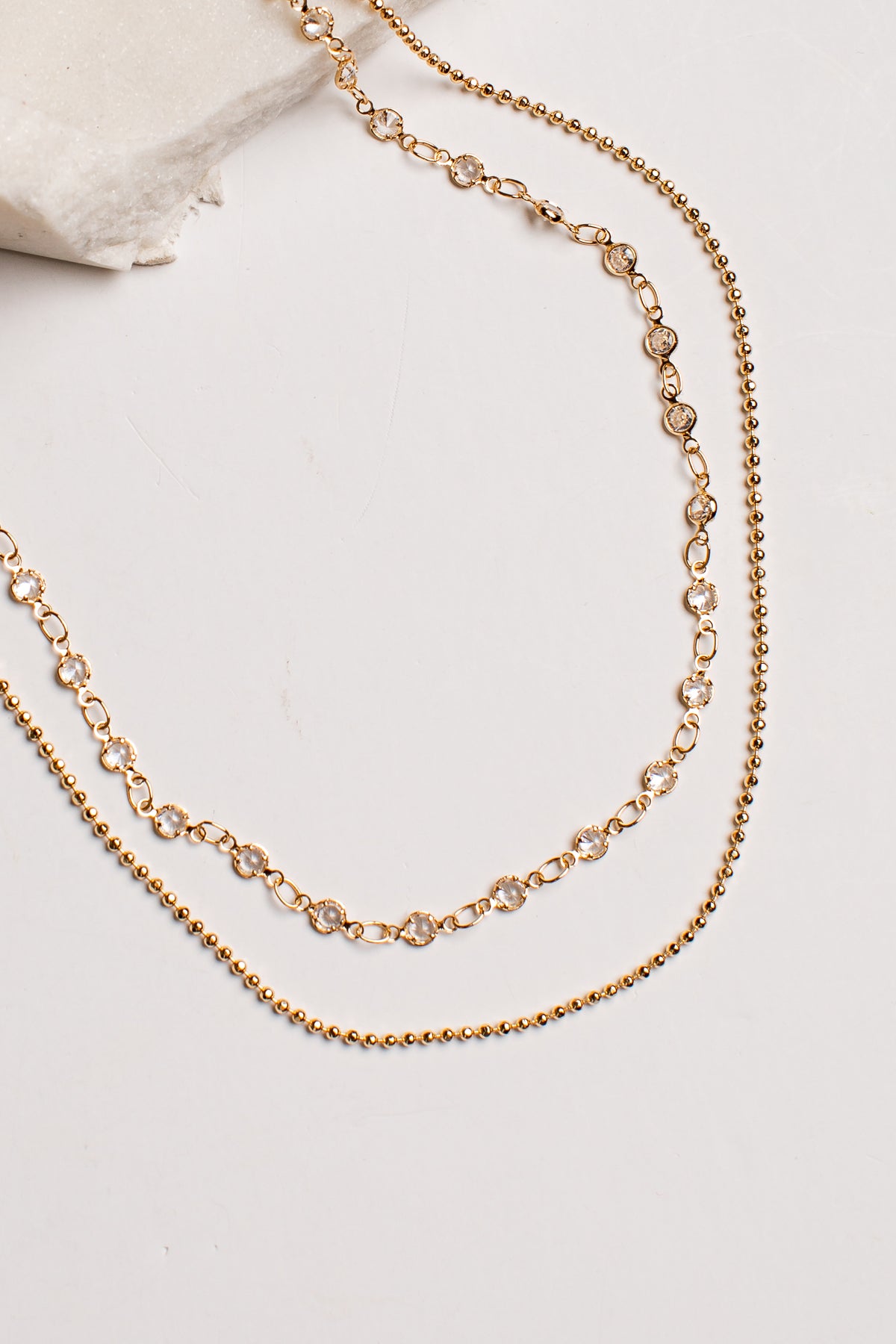 A double-layered gold chain necklace with one layer featuring small oval links and the other a finer beaded chain, displayed on a light neutral background.