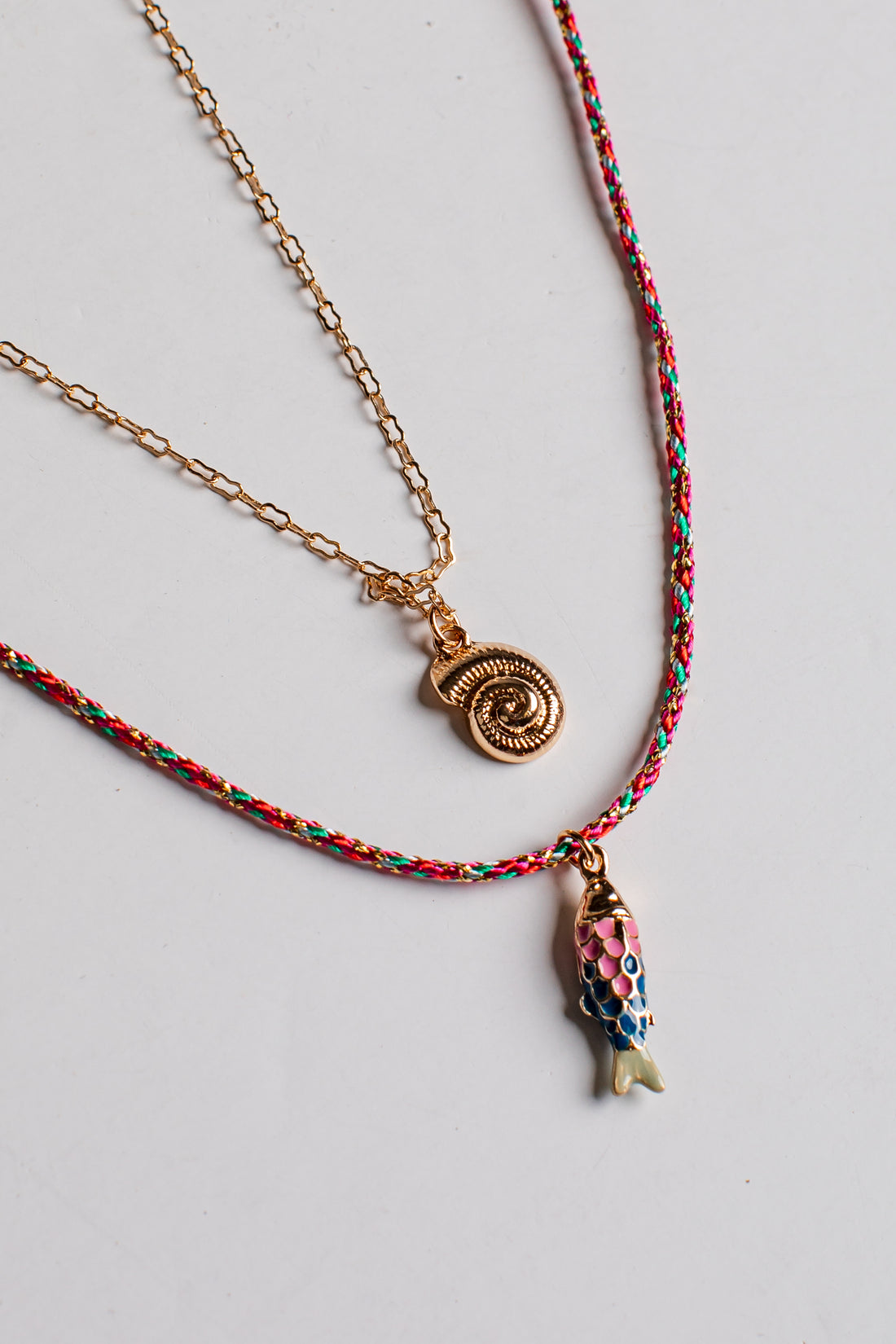 A layered necklace set on a light gray background. The top layer features a gold chain with a small gold spiral pendant.